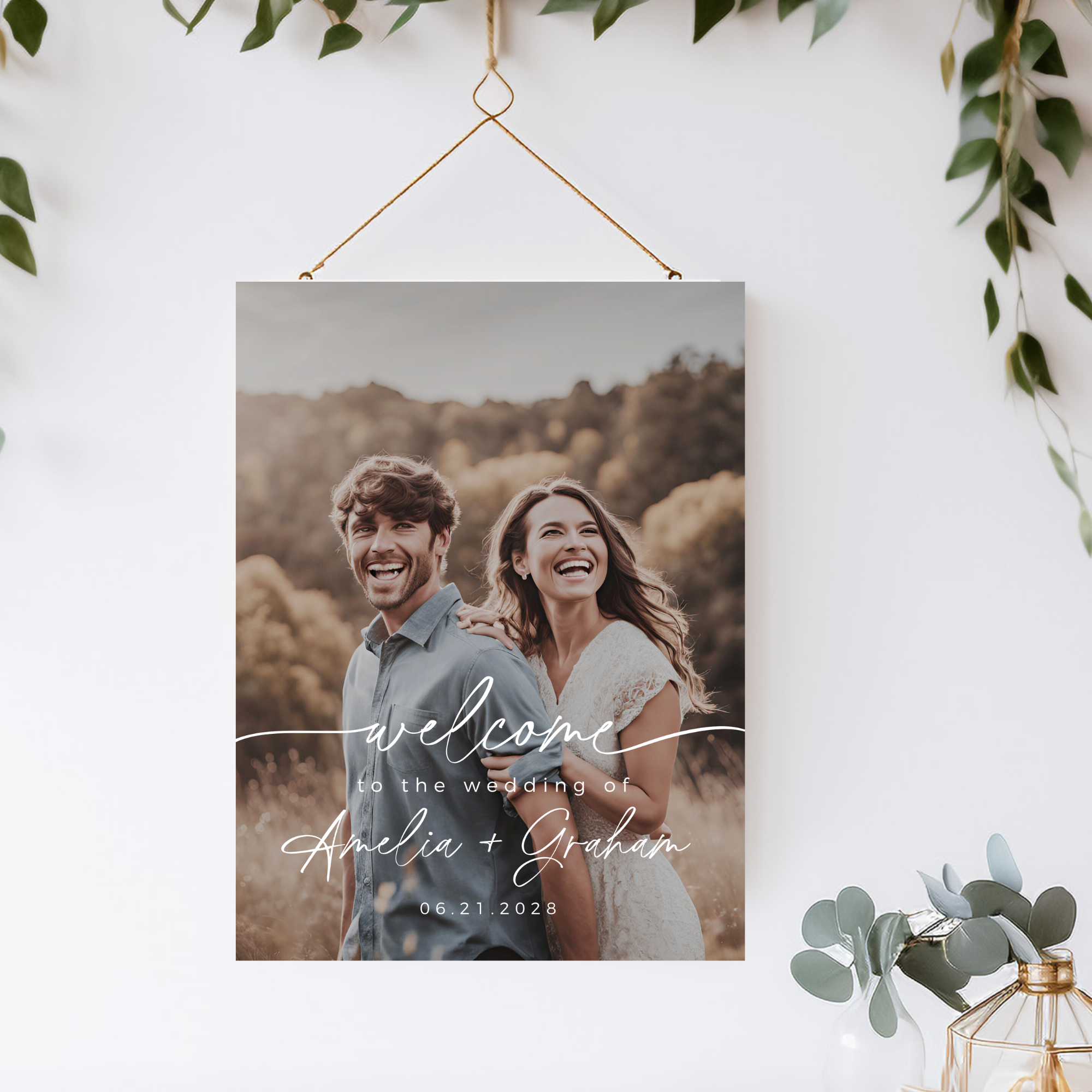 Wedding welcome sign with a couple in an outdoor setting, hanging on a wall with greenery.