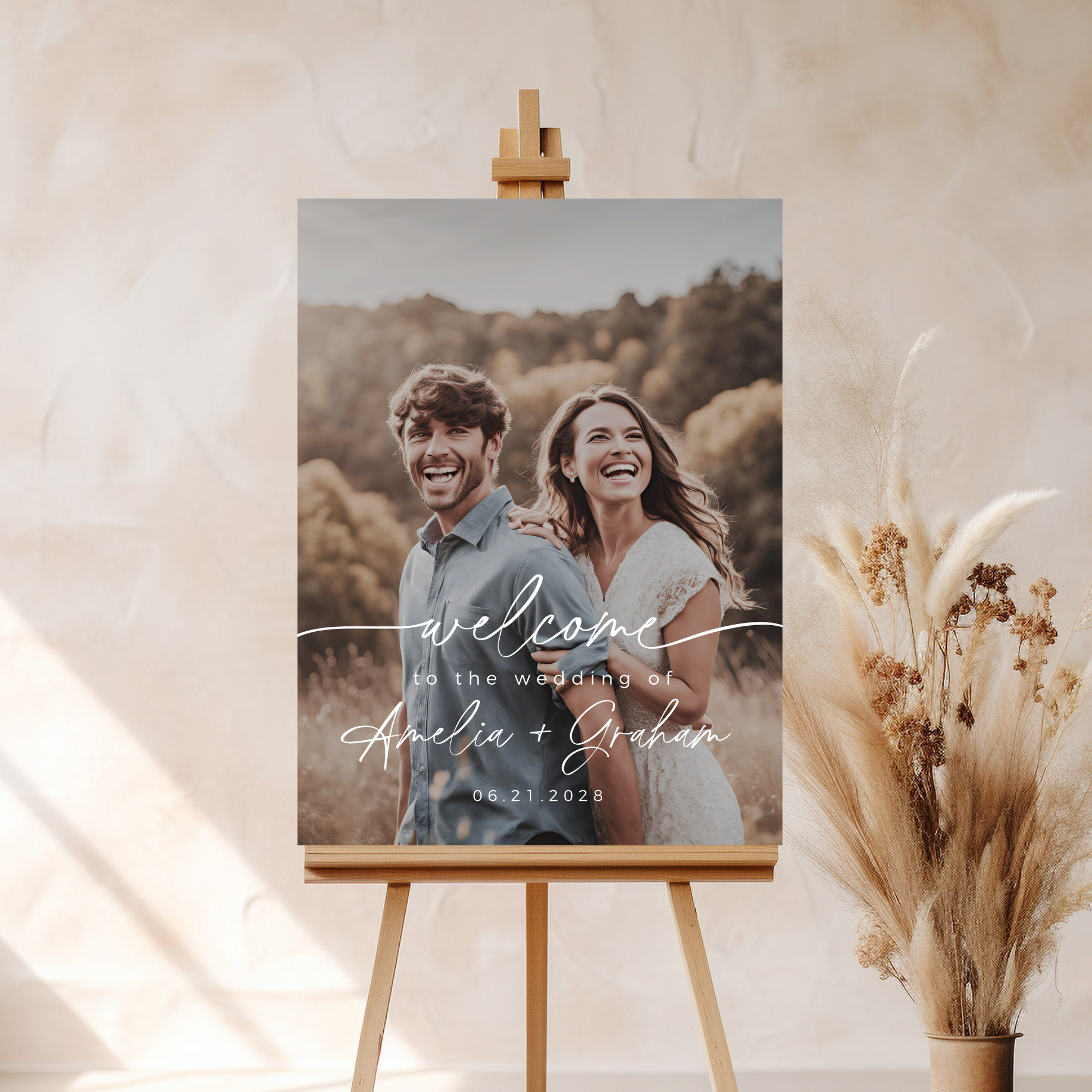 Wedding welcome sign with a couple's photo on an easel, surrounded by dried plants.